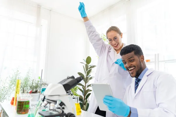 在实验室里与平板电脑打交道的年轻科学家男女们 他们愉快地讨论着成功的问题 并一起微笑着 显微镜和科学地坐在桌子上 — 图库照片