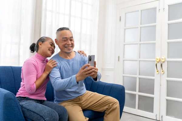 stock image Asian couple sitting on sofa in living room, They are use smartphone to selfie portrait them, smile with happiness together, copy space