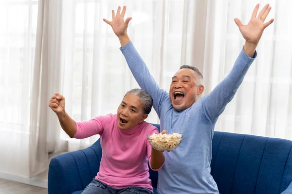 stock image Happily Asian Senior couple watching television and eating popcorn on sofa in living room at home. They are Raise hands above head glad that their team won. Enjoying leisure time. copy space