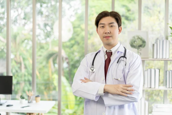 亚洲年轻男医生的画像 脖子上戴听诊器 双臂交叉 身穿白衣 笑着看着相机 专业医生的概念 — 图库照片