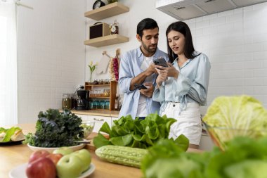 Uygulamaları paylaşmak için akıllı telefon kullanan genç çiftin portresi ya da mutlu mesut sosyal ağları kontrol eden evli, gülümseyen eşler evde mutfakta çevrimiçi cep telefonu oyunları oynuyorlar.