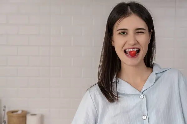 Portrait Young funny woman with a cherry fruit in her mouth standing in kitchen, smile and looking at camera, copy space
