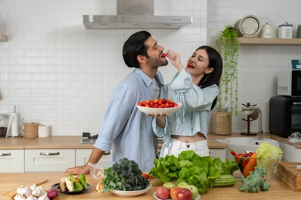 快乐快乐的年轻夫妇年轻漂亮的丈夫和漂亮的妻子在家里厨房一起品尝柑橘类水果 一起吃樱桃 健康的食物 饮食和爱情的概念 图库图片