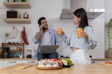 Mutlu Asyalı genç çift evdeki mutfakta birlikte yemek pişiriyor. Genç koca akıllı telefon ve dizüstü bilgisayar kullanırken güzel eş elinde bir bardak portakal suyu, aşk ve aile kavramı tutuyordu..