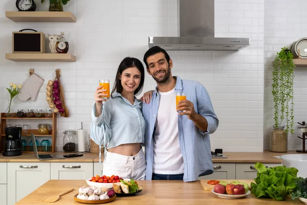 Happy asian young couple cooking together in the kitchen at home. Young husband use smartphone and laptop computer while beautiful wife holding glass of orange juice in hand, Love and family concept.