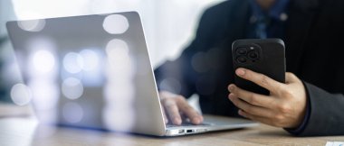 İşadamı masasında bir cep telefonu ve bir dizüstü bilgisayar kullanarak ofisindeki verimliliği artırıyor. Teknolojinin kusursuz bütünleşmesine tanık olun ve dijital çalışma ortamının faydalarını kucaklayın..