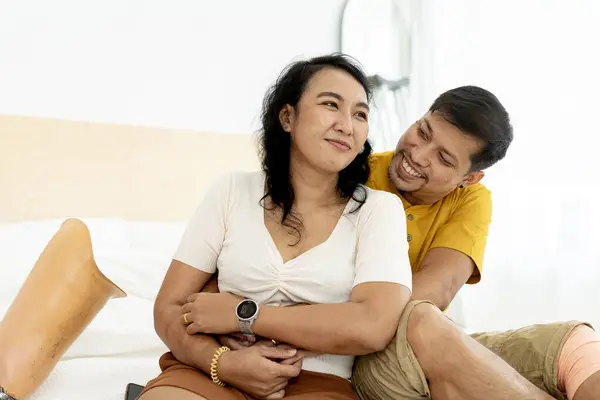 一位膝盖受伤的丈夫坐在床上 拥抱他残疾的妻子 她的假腿被摘掉了 一对快乐的亚洲夫妇在家里共度时光 图库照片