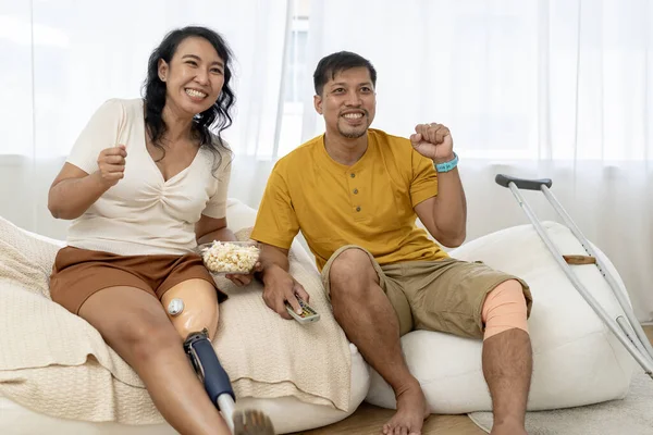 妻子戴着假腿 丈夫膝盖受伤 看电视 享受着在体育运动中欢呼的兴奋心情 一对快乐的亚洲夫妇在家里共度时光 图库图片