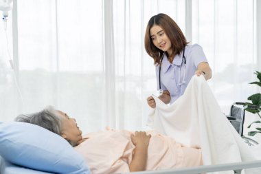 Yaşlı bir Asyalı hasta hastaneye yatırıldı. Yaşlı bir hastanın bakıcısı ya da hemşiresi yatakta yatan bir hastanın battaniyesini örtmeye yardım ediyor. Hastanelerde ya da kliniklerde hastalara hemşireler bakar.