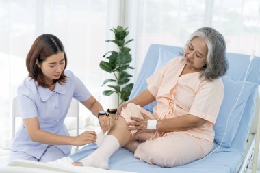 Yaşlı Asyalı hasta hastaneye yatırıldı. Hemşire, hastanın yaralı bacağındaki bandajla ilgilenir. Hemşire, hastane ya da klinikte hastalara bakar..