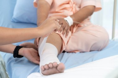 Hastaneye yatırılan yaşlı bir Asyalı hastanın yakın plan fotoğrafı. Bir hemşire yaralı bir hastanın bacağındaki bandajla ilgilenir. Hemşire, hastane ya da klinikte hastalara bakar..