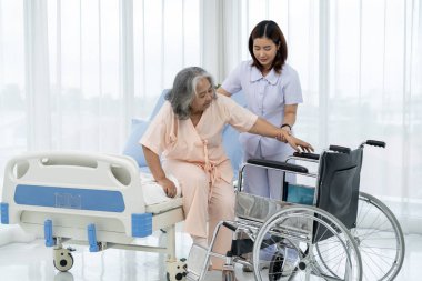 Bir hemşire, yaşlı bir Asyalı hastayı bir kazada bacağının röntgenini çektirmek için tekerlekli sandalyeye taşıyor. Hemşire, hastane ya da klinikte hastalara bakar..