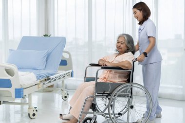 Bir hemşire yaşlı bir Asyalı hastayı tekerlekli sandalyeye oturtup bacağının bir kazada yaralandığı röntgenini çektirmeye götürüyor. Hemşire, hastane ya da klinikte hastalara bakar..