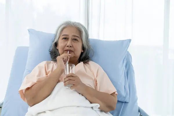 Elderly Asian patient admitted to hospital During her recovery, The patient was feeling thirsty and drinking water while lying on the bed. Nurses care for patients in hospitals or clinics.