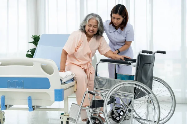 A nurse carries an elderly Asian patient into a wheelchair to take him to get an x-ray of his leg injured in an accident. A nurse takes care of patients in a hospital or clinic.