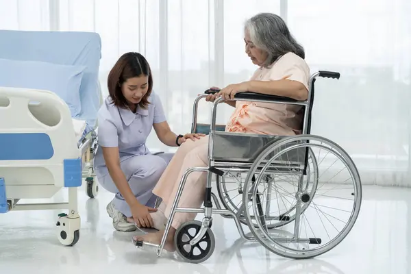 A nurse loads an elderly Asian patient into a wheelchair and makes a footrest ready to take him to get an x-ray of his leg injured in an accident. A nurse takes care of patients in a hospital