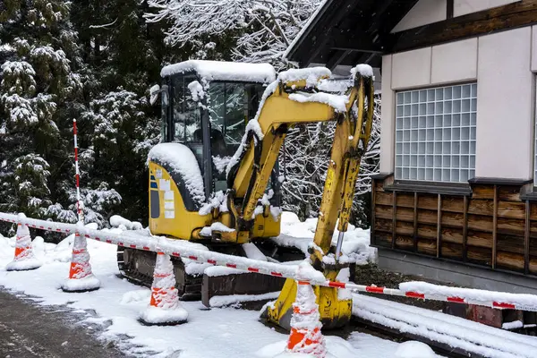 Yellow excavator covered in snow next to building at construction site. Perfect for winter construction, snow removal, and construction equipment photos.