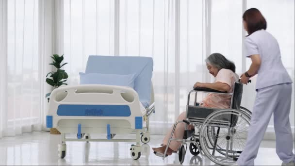 ハンドヘルドショット 看護師は 事故で負傷した足のX線のために彼を取った後 車椅子からベッドにアジアの高齢の患者を運びます 看護師が病院で患者の世話をしています — ストック動画