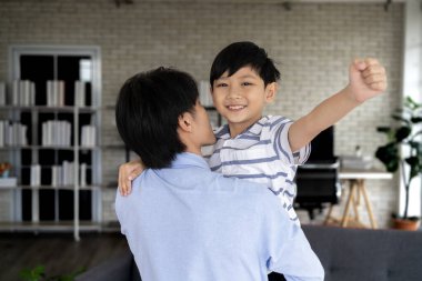 Mutlu Asyalı baba, küçük oğluna evinde, oturma odasında sarılıyor. Bir baba, mutlu bir şekilde gülümseyen ve kollarını sevinçle kaldıran oğluna sarılır. Aile mutluluğu oturma odasında.