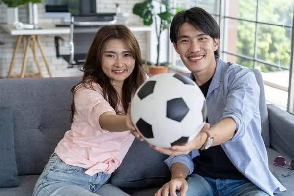 Couple holding a soccer ball and cheering football at home. Young Asian couple watching soccer match on tv and cheering football team at television in living room, sports and home entertainment