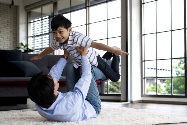 快乐的年轻亚洲父亲躺在客厅地板上 和儿子一起玩飞机 他在家里抱着兴奋而快乐的小儿子 客厅里的家庭幸福 图库图片