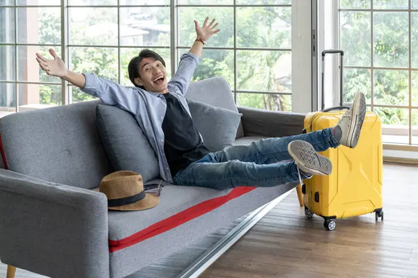 Happy man returning home from summer vacation trip Sitting on the sofa in the living room with a yellow suitcase. Joyful man feeling excited returning home after vacation travel.