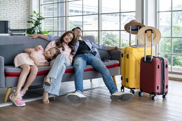 Tired couple and their daughter return home from a summer vacation trip. Sitting asleep on the sofa in the living room with a yellow and red suitcase. Returning from adventure and vacation in holiday