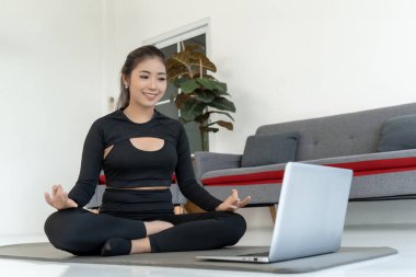 Spor giyim bölümünde genç bir Asyalı kadın evde yoga yapıyor. Bir dizüstü bilgisayarın önünde yoga minderinin üzerinde oturmuş meditasyon yapıyor ve esniyor. Bu görüntü fitness, sağlık ve yaşam tarzı blogları ve makaleleri için mükemmeldir.