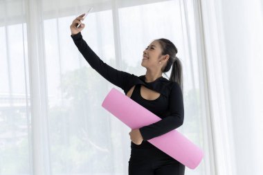 Gülümseyen Asyalı bir kadın yoga minderini tutuyor ve yoga stüdyosunda selfie çekiyor. Sportif kıyafetler giyiyor ve mutlu ve sağlıklı görünüyor.. 
