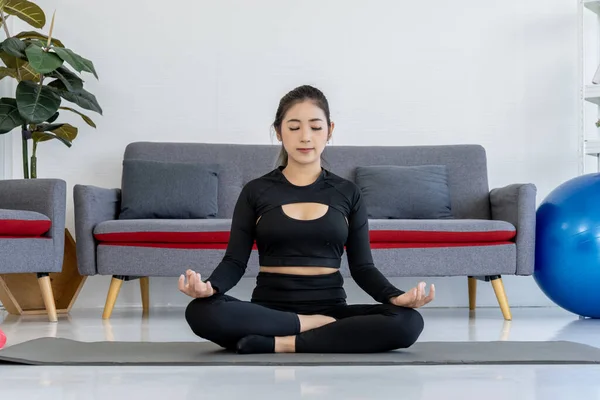 A young Asian woman in sportswear practices yoga at home, meditating on a yoga mat in her living room. Serene expression on her face, she finds inner peace and balance through yoga meditation.