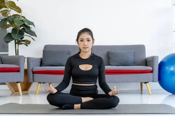 A young Asian woman practices yoga in her living room, showcasing a healthy lifestyle. She sits in the lotus pose on her yoga mat, demonstrating a simple beginner yoga pose.