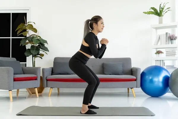 Asian woman squatting on yoga mat in living room, exercising her legs and glutes. Healthy lifestyle concept with home workout.