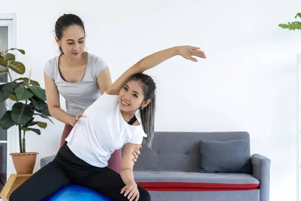 Personal trainer is assisting a client with a stretch on a fitness ball in a gym setting. This image is perfect for marketing materials for personal trainers and fitness studios.