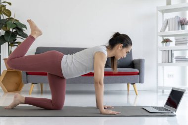 Genç bir kadın evindeki oturma odasında dizüstü bilgisayarından yoga pozları öğreniyor. Gülümsüyor ve eğleniyor. Formu da iyi..