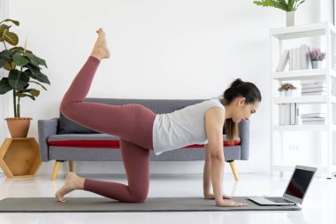 Genç bir kadın evindeki oturma odasında dizüstü bilgisayarından yoga pozları öğreniyor. Gülümsüyor ve eğleniyor. Formu da iyi..