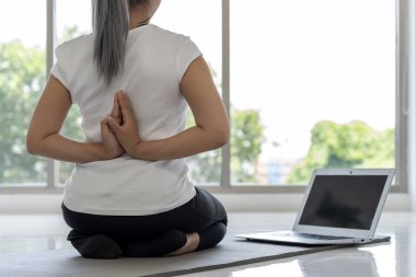 Genç bir kadın evindeki oturma odasında dizüstü bilgisayarından yoga pozları öğreniyor. Gülümsüyor ve eğleniyor. Formu da iyi..