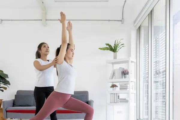 Asian yoga teacher demonstrates stretching pose while guiding her student towards proper alignment technique. Tranquil setting and focused expressions convey essence of yoga's transformative power.