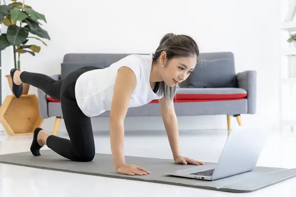 A young woman learns yoga poses from an online tutorial on her laptop in her living room at home. She is smiling and enjoying herself, and her form is good.