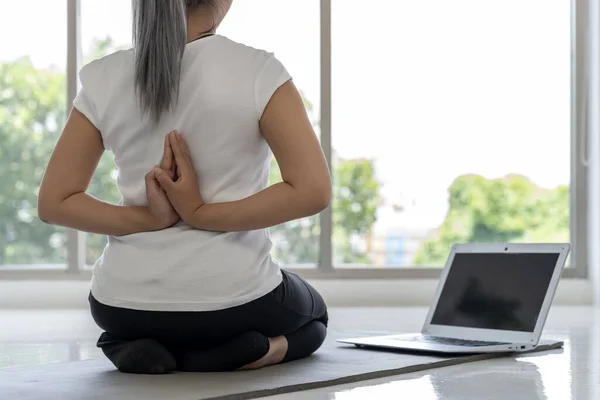 A young woman learns yoga poses from an online tutorial on her laptop in her living room at home. She is smiling and enjoying herself, and her form is good.