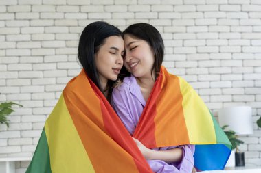 İki gülümseyen genç kadın gururla vücutlarını bir yatak odasında gökkuşağı bayrağıyla örterek LGBTQ gururunu gösteriyor ve çeşitliliği kutluyorlar. Sevgi, kabullenme ve özgürlük hareketine katılın