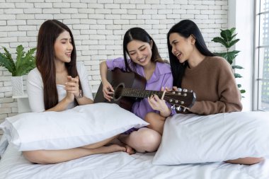 Kadınlar bir yatakta gitarla şarkı söyleyip gülüyorlar. Harika zaman geçiriyorlar ve müzik bulaşıcı..