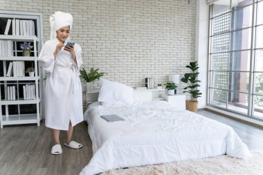 Yaşam tarzı, beyaz bir bornoza sarılı ıslak saçlı genç bir bayanın akıllı telefon cihazıyla konuşurken yatak odasında duruşu..