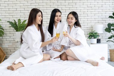 Üç genç kadın bir yatakta oturmuş şık beyaz bornozlarla bardaklarını tokuştururken coşkulu bir kutlama. Yatak odasında neşeli bir partinin tadını çıkarmak, neşe ve beraberlik dolu neşeli bir atmosfer yaratmak.