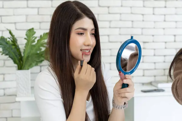 Young female with black hair puts on lip makeup in bedroom while viewing reflection in a round mirror on bed, lifestyle scene of young adults applying cosmetics