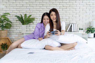 Teknoloji zevki, yatakta iki genç kadın, yatak odasında akıllı telefon oyunu oynuyorlar. Bu rahat ortamın dijital eğlencesine dalın, modern boş zamanların özünü yakalayın