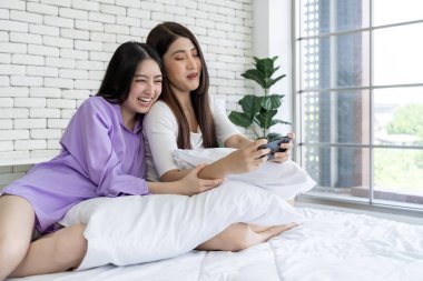 Teknoloji zevki, yatakta iki genç kadın, yatak odasında akıllı telefon oyunu oynuyorlar. Bu rahat ortamın dijital eğlencesine dalın, modern boş zamanların özünü yakalayın