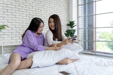 Teknoloji zevki, yatakta iki genç kadın, yatak odasında akıllı telefon oyunu oynuyorlar. Bu rahat ortamın dijital eğlencesine dalın, modern boş zamanların özünü yakalayın