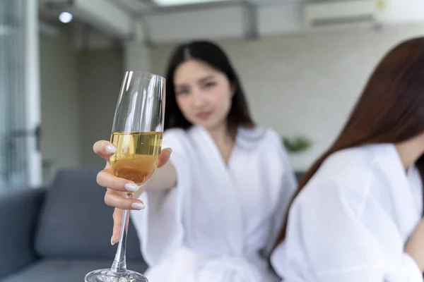 Casual indoor scene of a smiling black haired woman dressed in a white robe holding up a wine glass, lifestyle photo promoting relaxation and home events.