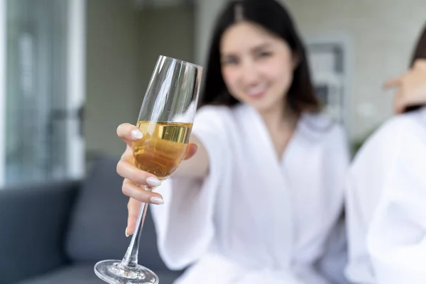 Casual indoor scene of a smiling black haired woman dressed in a white robe holding up a wine glass, lifestyle photo promoting relaxation and home events.