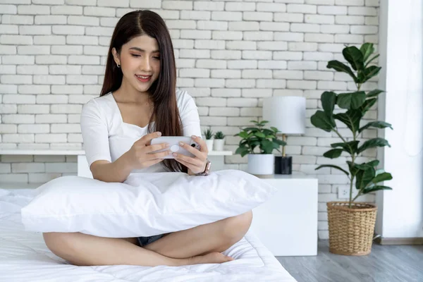 A casual day of a young woman on a bed in a bedroom. A young woman sitting with pillows on a bed and play game on smartphone